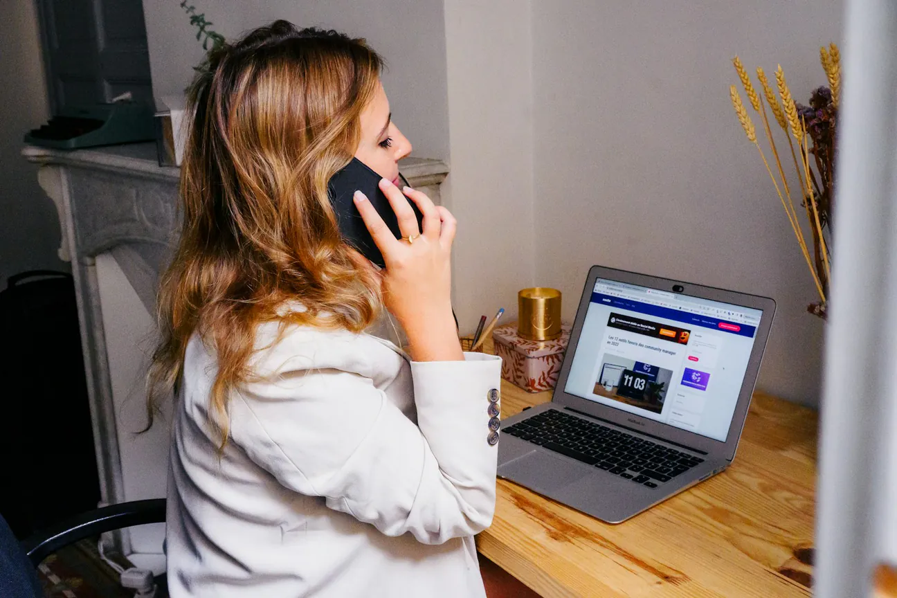 A woman at a wooden desk taking a call on her mobile while an open laptop shows the dashboard she is working in