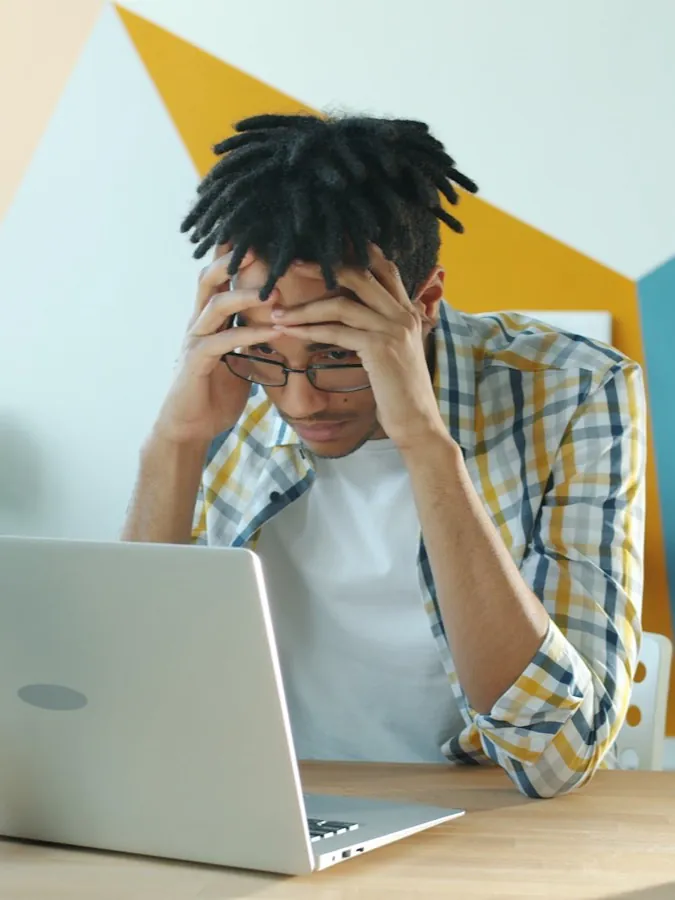 A man at a desk with his head in both hands looking down at an open laptop beside a cooling mug of coffee