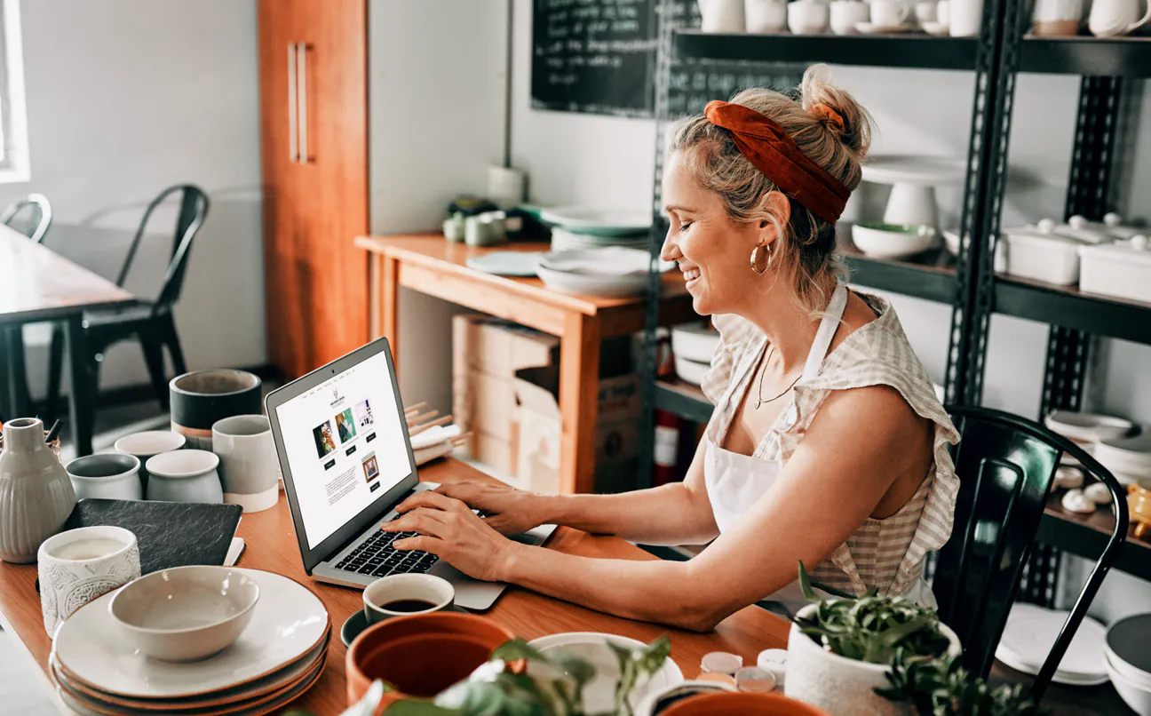 Shop owner in an apron works on a web page on her laptop among ceramics