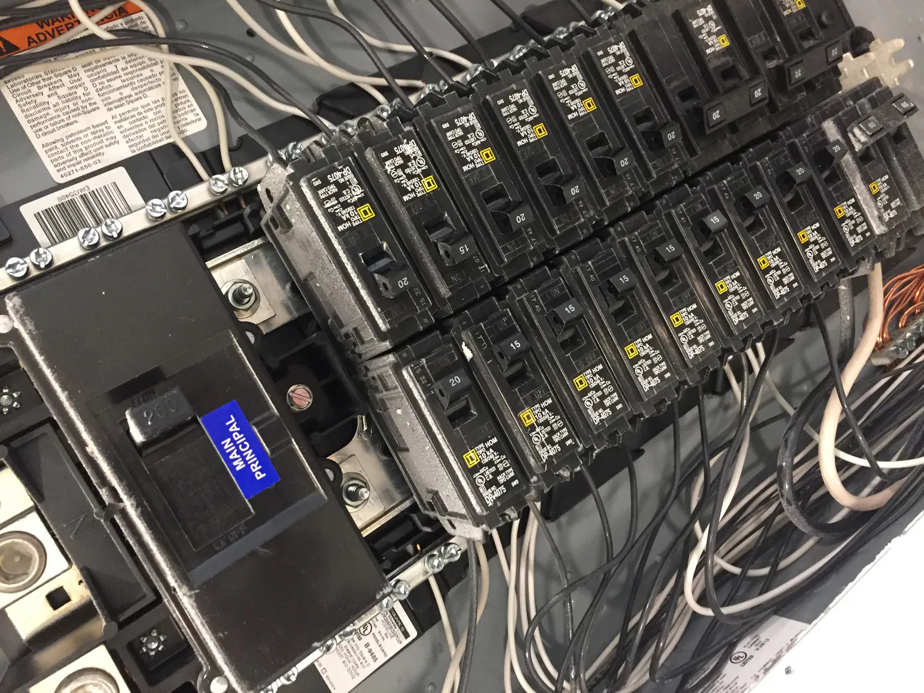 The inside of a residential breaker panel with its cover off, the main breaker labeled at the left and two rows of branch breakers with white and black conductors landing on the neutral and ground bars