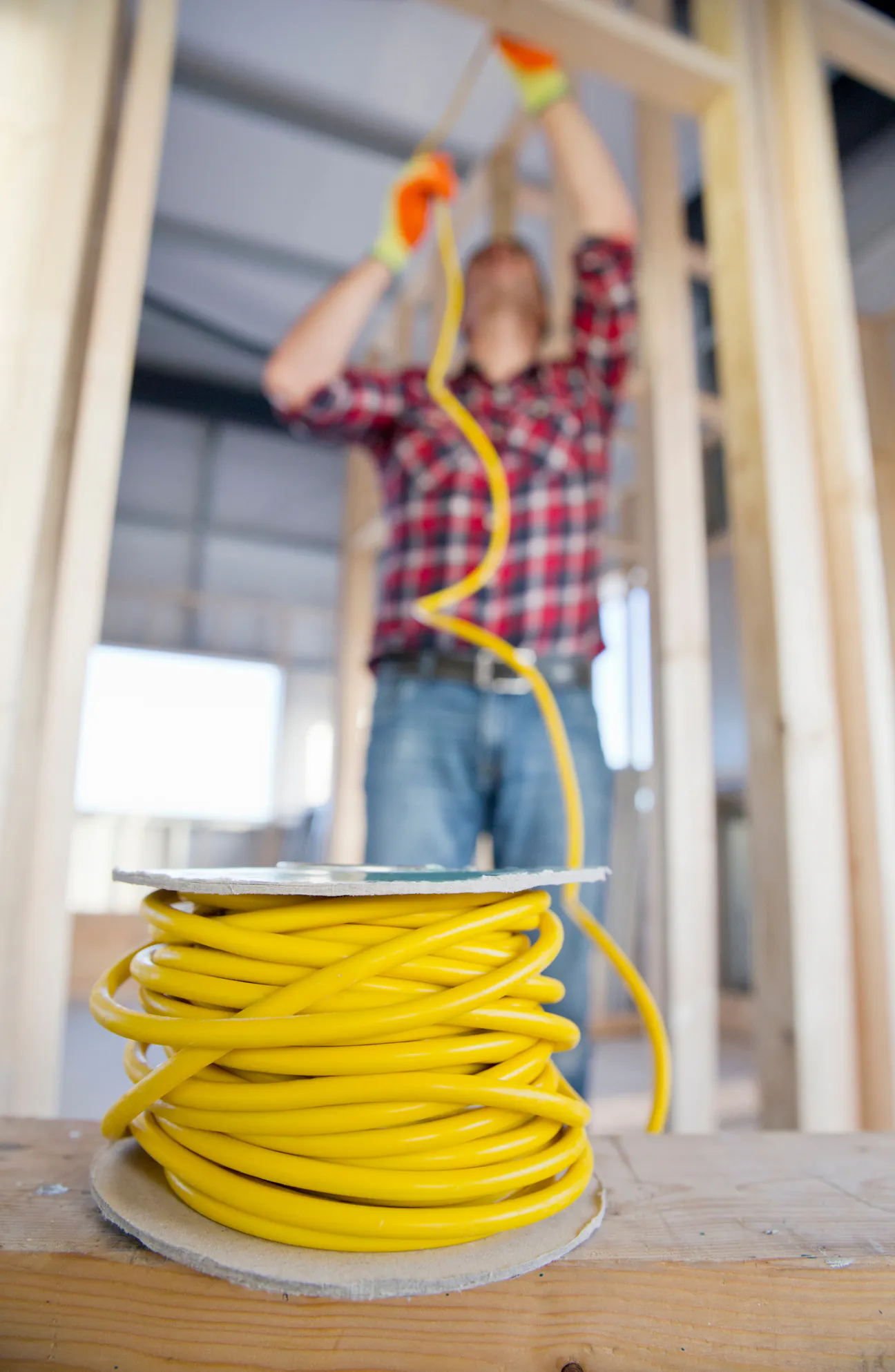 A large spool of yellow electrical cable on a timber sill in the foreground, with an electrician in a plaid shirt and work gloves out of focus behind it, running the same cable overhead through the open stud framing of an unfinished building