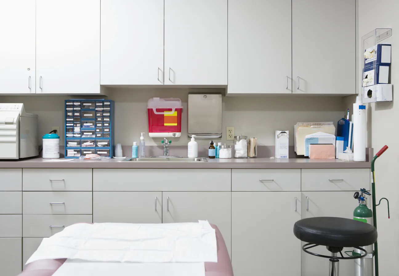 An exam room counter run with a small autoclave, a blue parts cabinet, a sink with soap and hand sanitizer, a sharps container and a paper towel dispenser, a paper covered exam table and a rolling stool in the foreground
