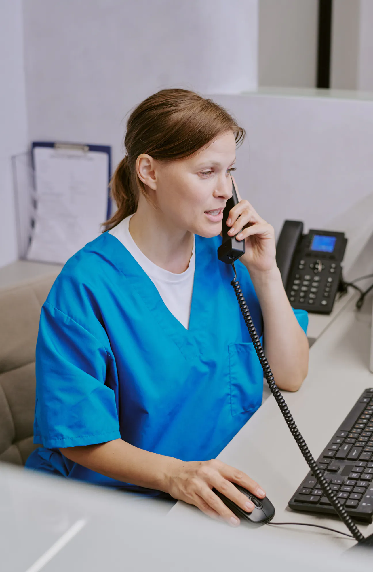 A medical receptionist in blue scrubs at the practice front desk, corded phone to her ear mid-call, her other hand at the keyboard, a clipboard of forms racked behind her