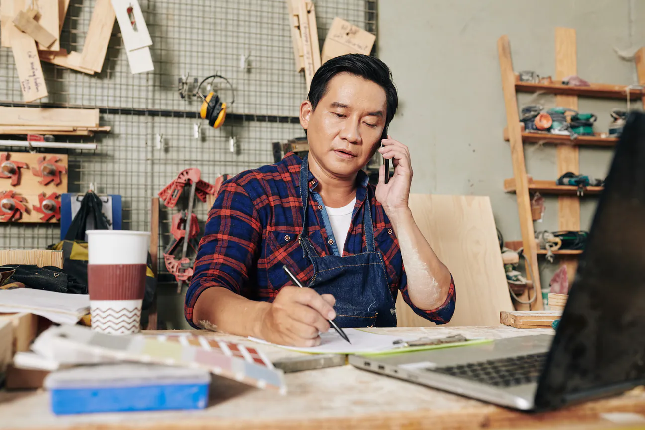 A workshop owner in a plaid shirt and denim apron sitting at his bench desk with the phone to his ear, writing on a work order beside a laptop and a color swatch card
