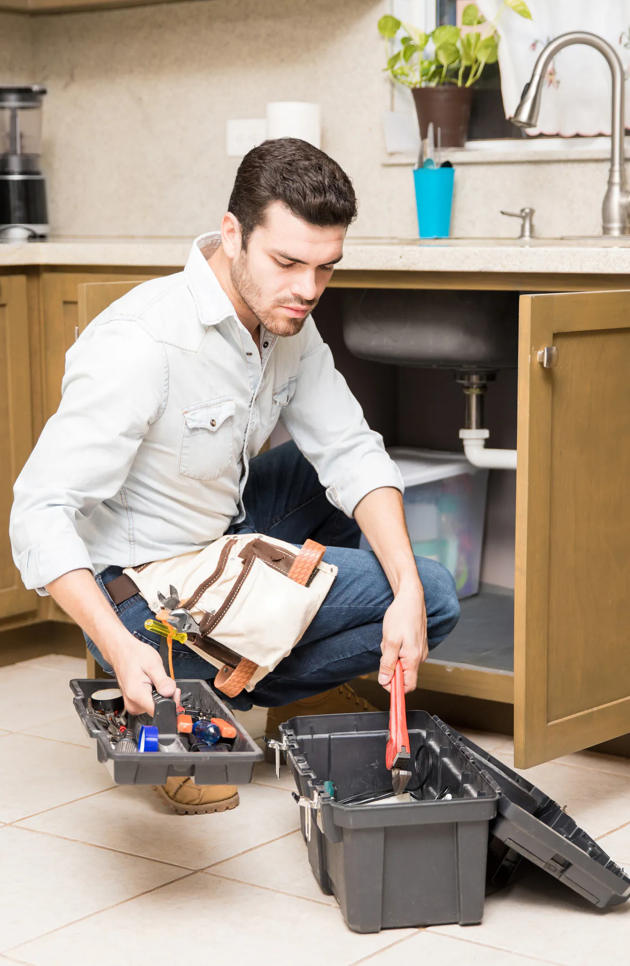 A handyman in a light denim work shirt with a canvas tool pouch on his belt, crouched on a tiled kitchen floor in front of an open sink cabinet, lifting a red handled pipe wrench out of a black tool tote while holding the lift out parts tray in his other hand