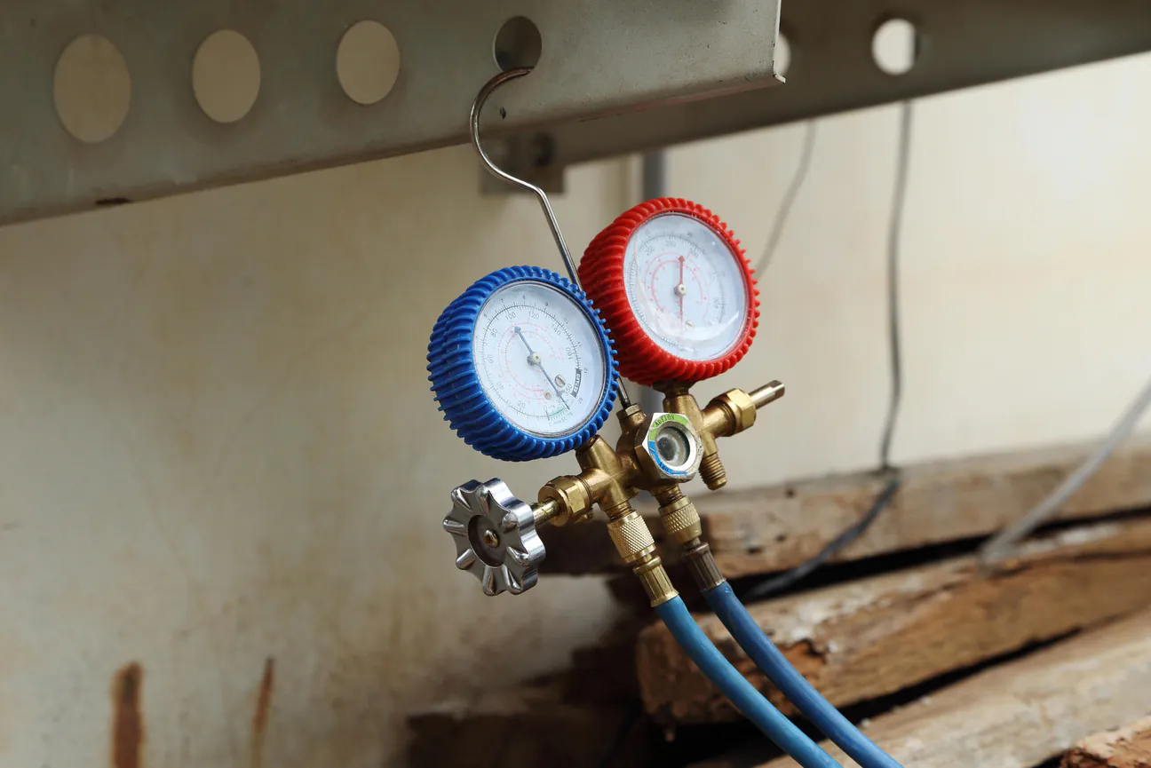A refrigerant manifold gauge set with blue and red gauges and brass body, hooked over a steel beam with its charging hoses hanging down, in front of a weathered wall