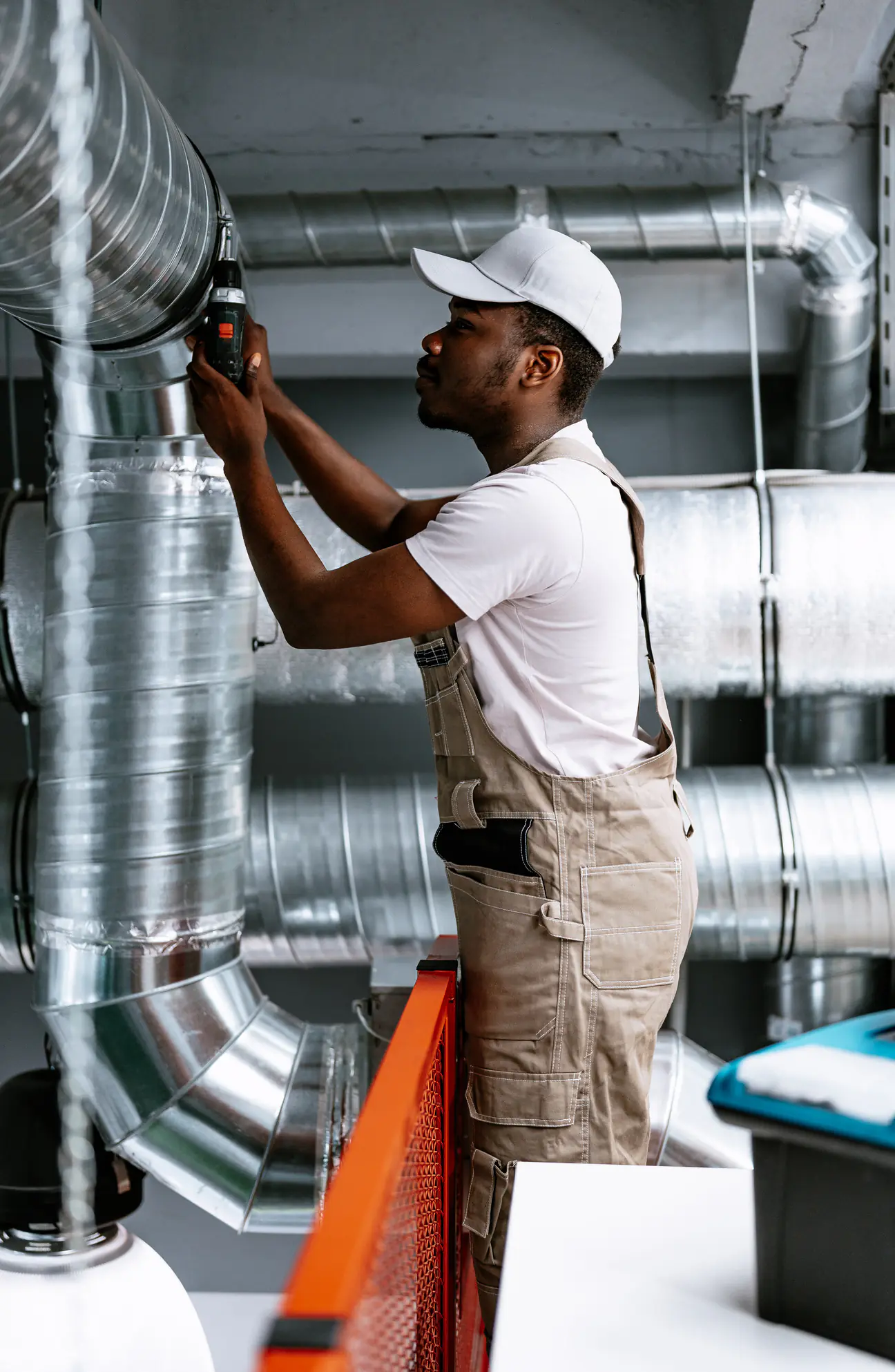 A technician in a white cap and tan overalls standing on a step ladder in a commercial mechanical space, drilling into a large spiral duct with more spiral ductwork running behind him