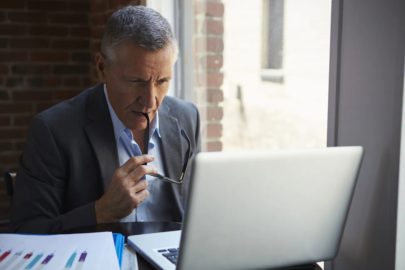 A gray haired man in a suit at a desk beside a tall office window, glasses held in one hand, reading a laptop screen with a printed chart on the desk in front of him