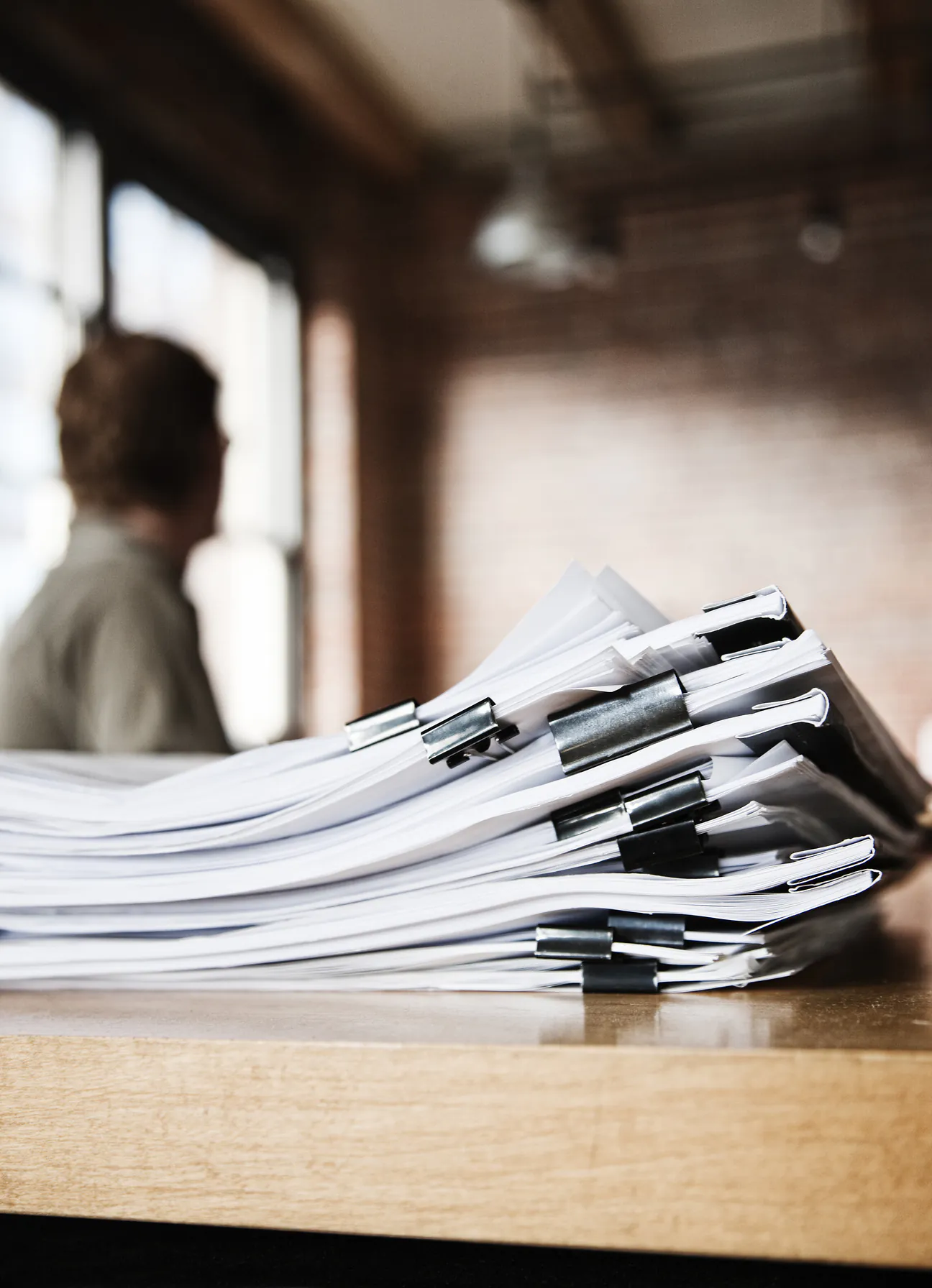 A tall stack of clipped document bundles on a wooden counter in a brick walled office, with a person out of focus at a window behind them