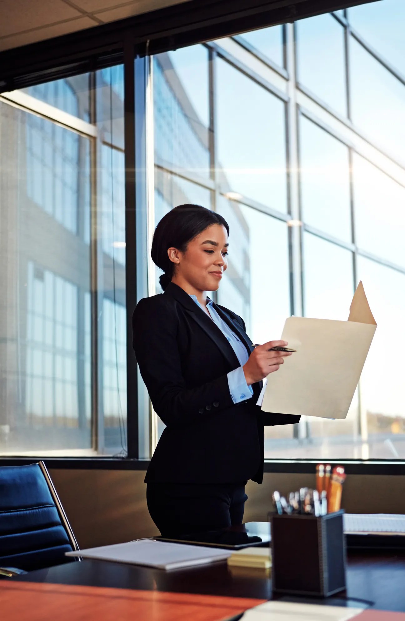 A lawyer in a black suit and light blue shirt standing at a dark wood desk beside a floor to ceiling office window, reading an open kraft file folder with a pen in her hand, with a bound document, a yellow legal pad and a pen cup on the desk in front of her