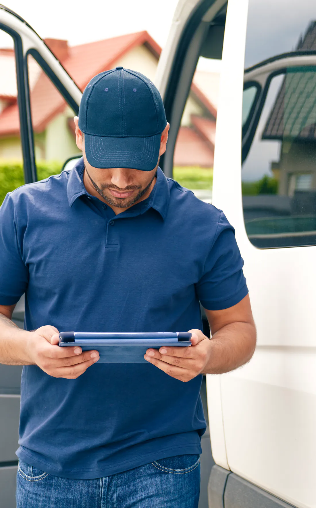 A plumbing company owner in a ball cap and navy polo standing at the open door of his white service van in a suburban driveway, reading a tablet