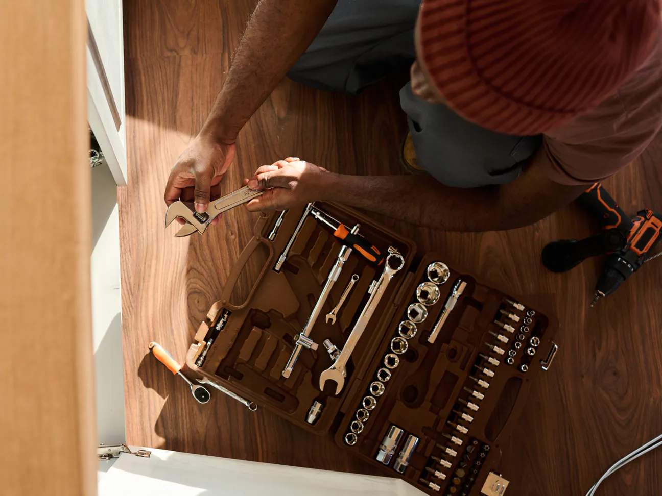 Overhead view of a plumber's hands holding an adjustable wrench over an open socket set on a warm walnut floor, beside an opened sink cabinet