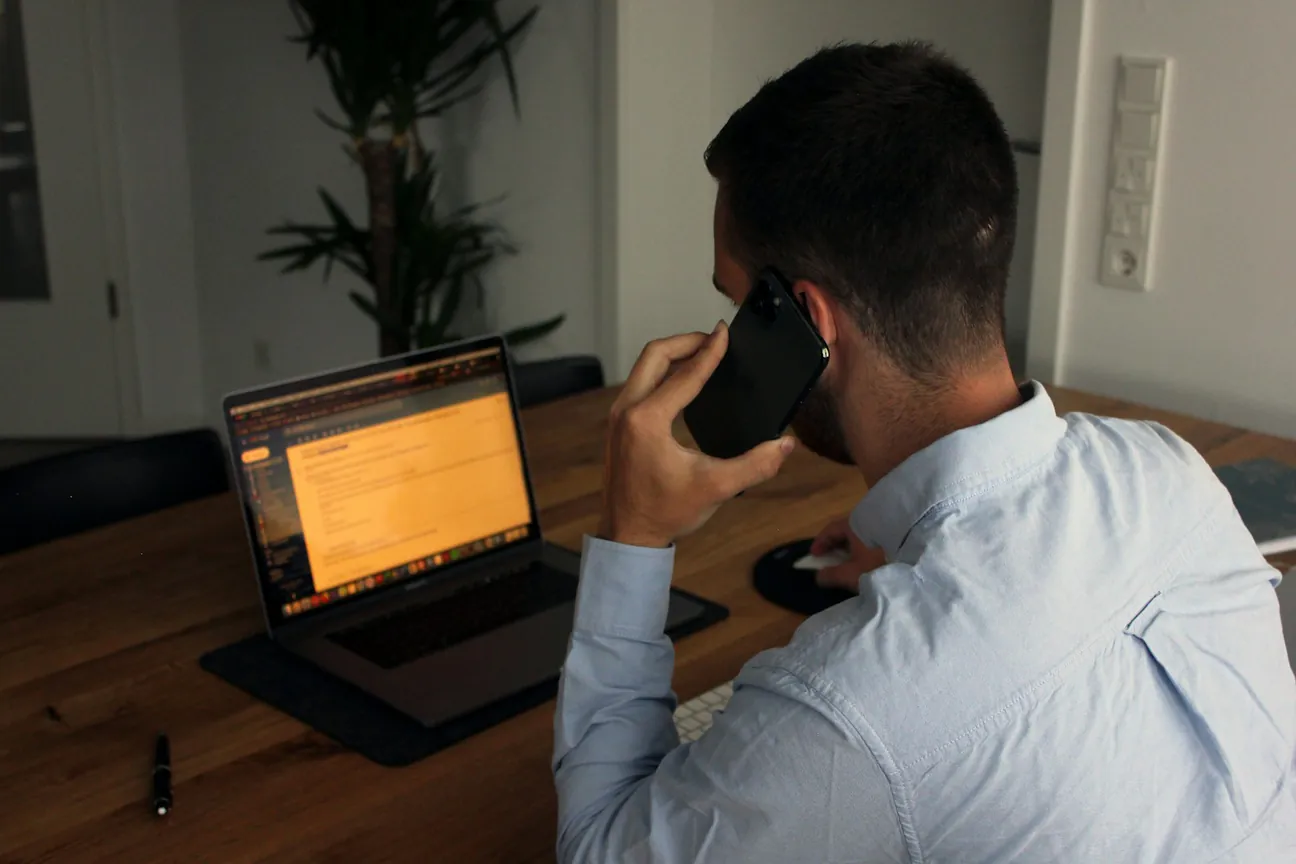 A man in a light blue shirt at a wooden desk holding a phone to his ear while an open laptop shows the document he is talking through