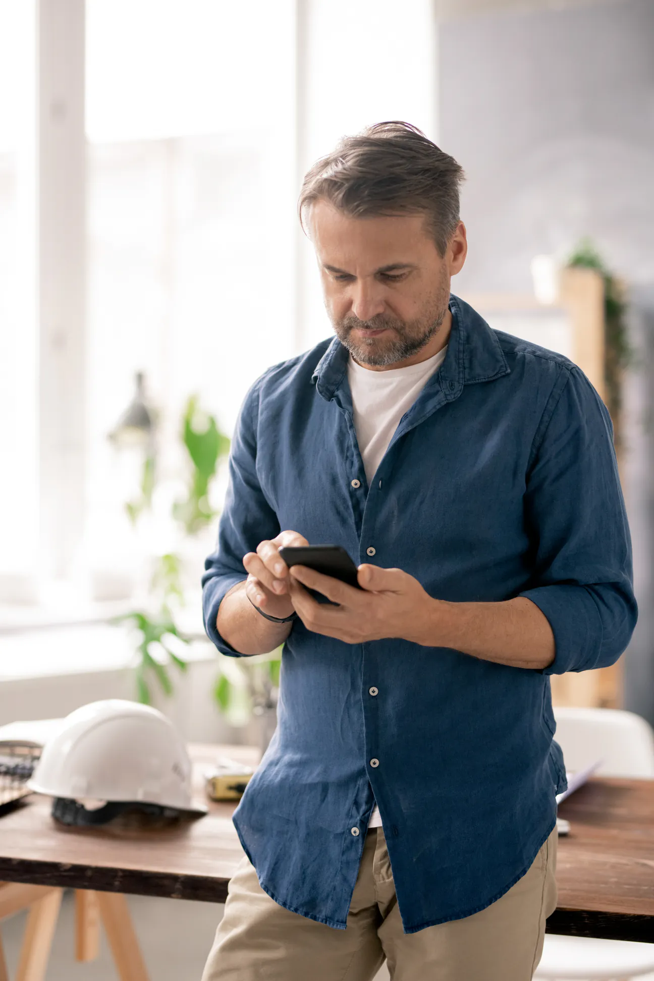 A contractor standing at his workbench scrolling his phone, hard hat on the desk beside him