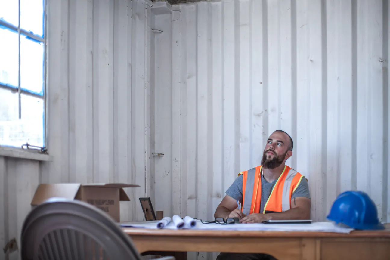 A bearded contractor in an orange hi-vis vest sitting at a wooden desk inside a corrugated metal jobsite cabin, rolled blueprints, reading glasses and a blue hard hat on the desk in front of him, looking up in thought