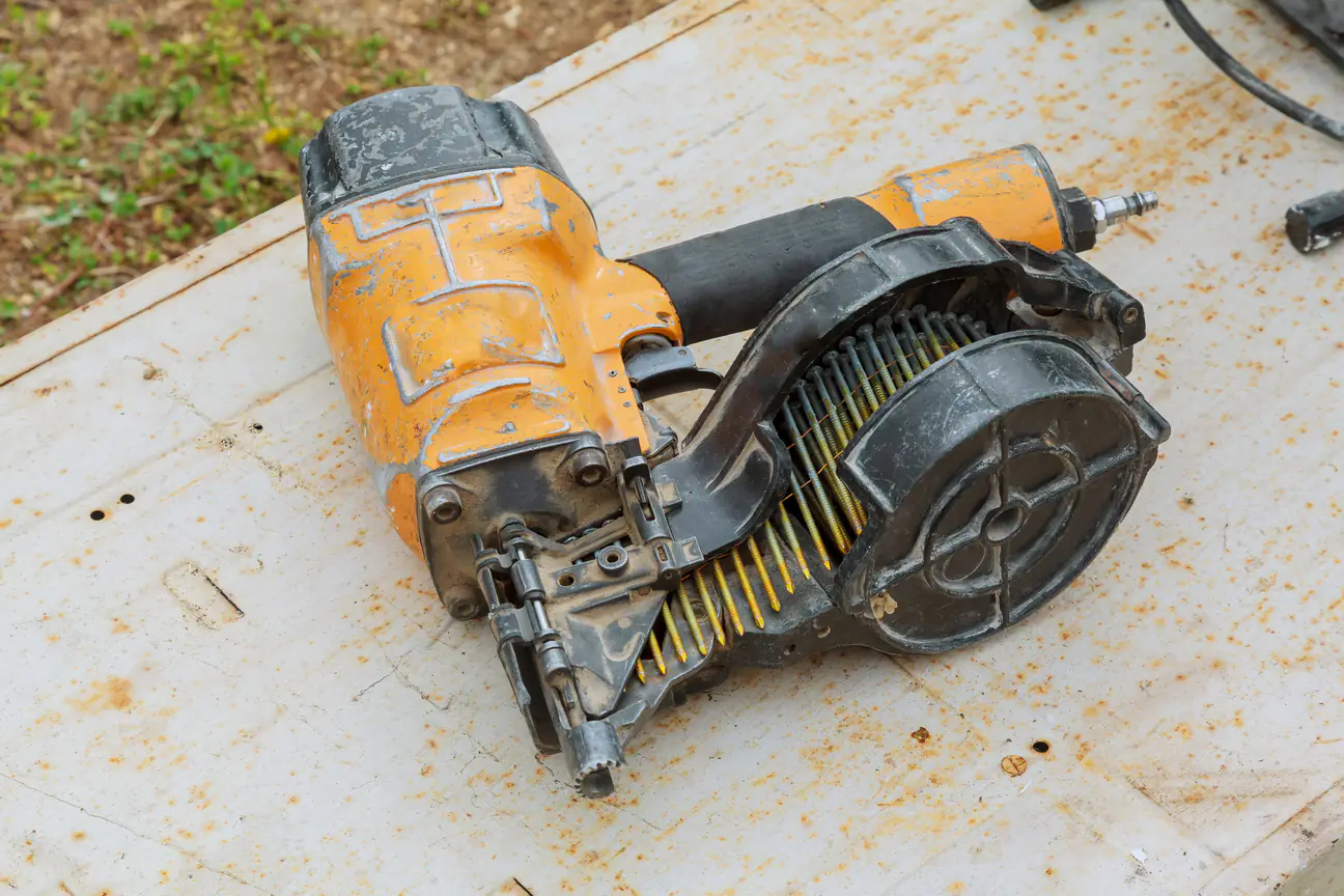 A well used orange and black pneumatic coil roofing nailer loaded with a coil of nails, lying on a sheet of weathered plywood sheathing beside a patch of grass