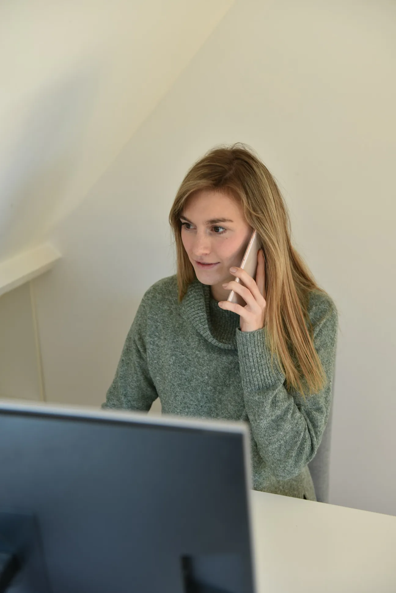 A woman in a grey-green sweater at her desk answering a call on her mobile with a monitor in front of her