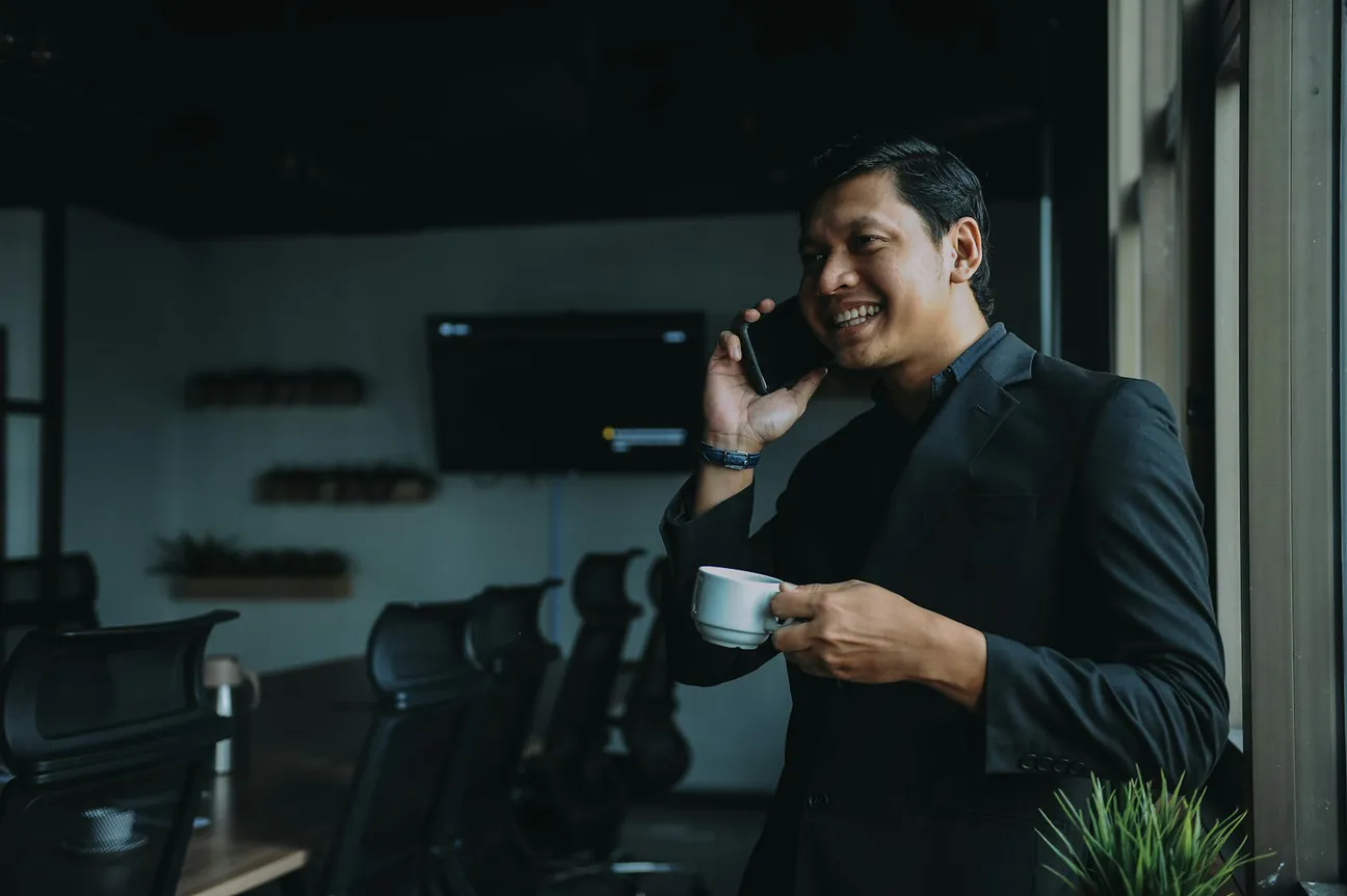 A smiling man in a dark blazer standing by a window with a coffee cup, taking a call on his mobile device in a quiet meeting room