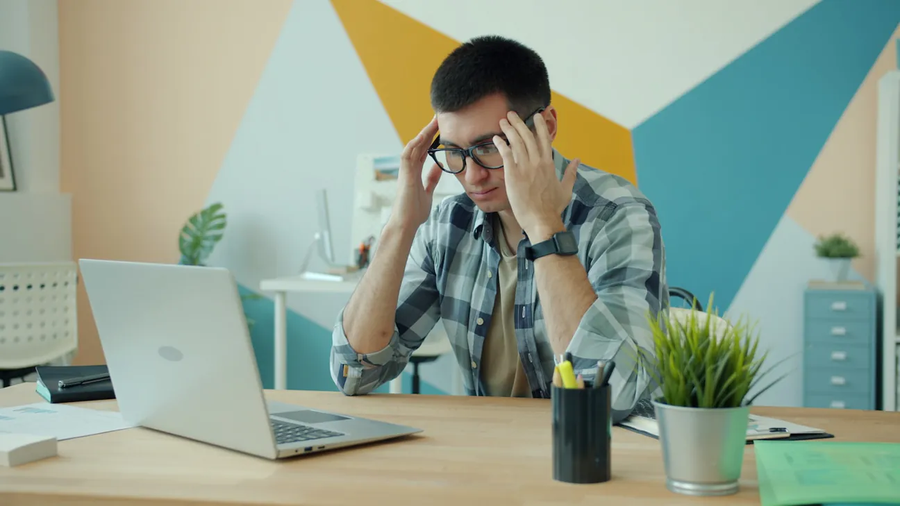 A man in a checked shirt with his hands at his temples staring down at an open laptop on a bright office desk