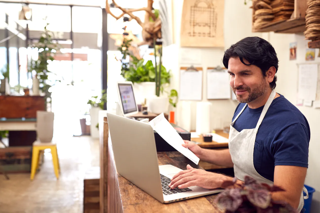 Shop owner in an apron working on a laptop at the front counter of his store