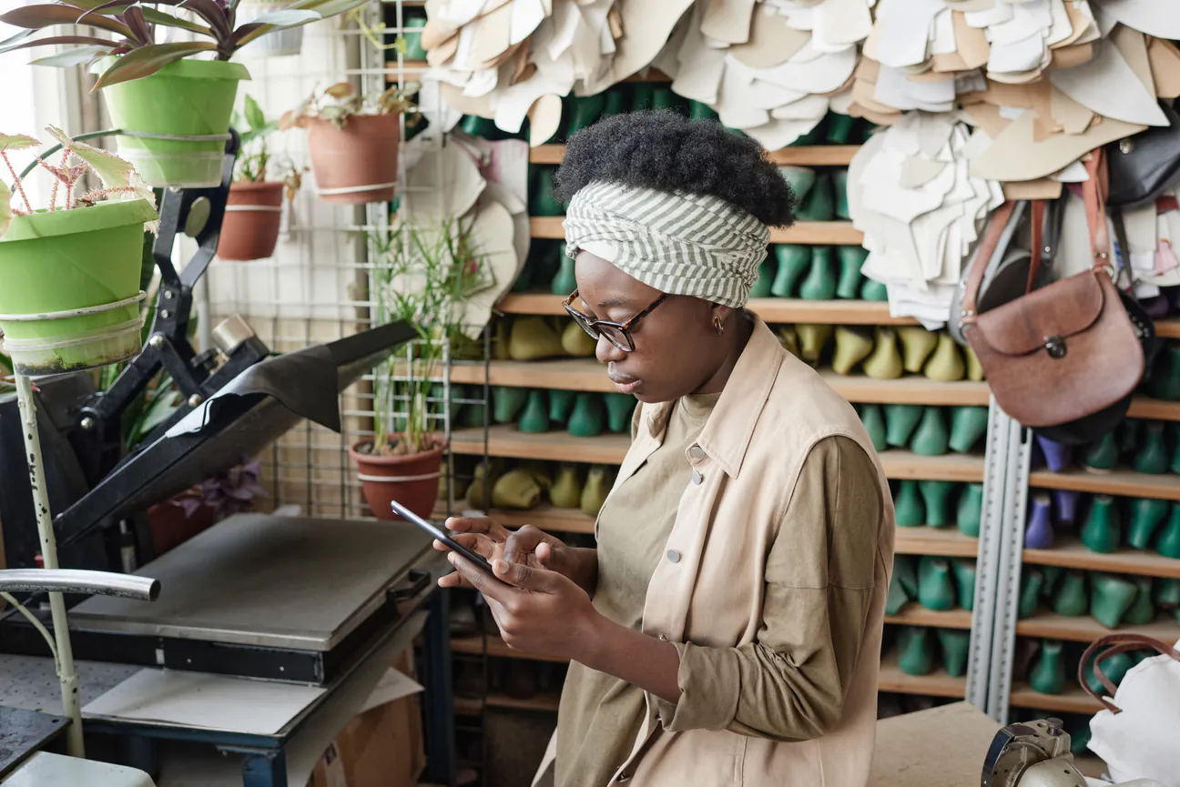 Shoemaker in glasses and a headwrap types a question on her tablet in her workshop