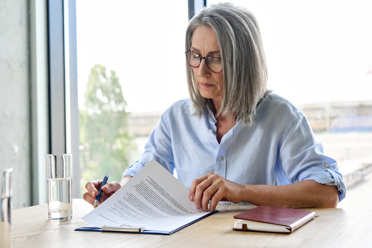Woman in glasses reads a printed agreement line by line, pen in hand
