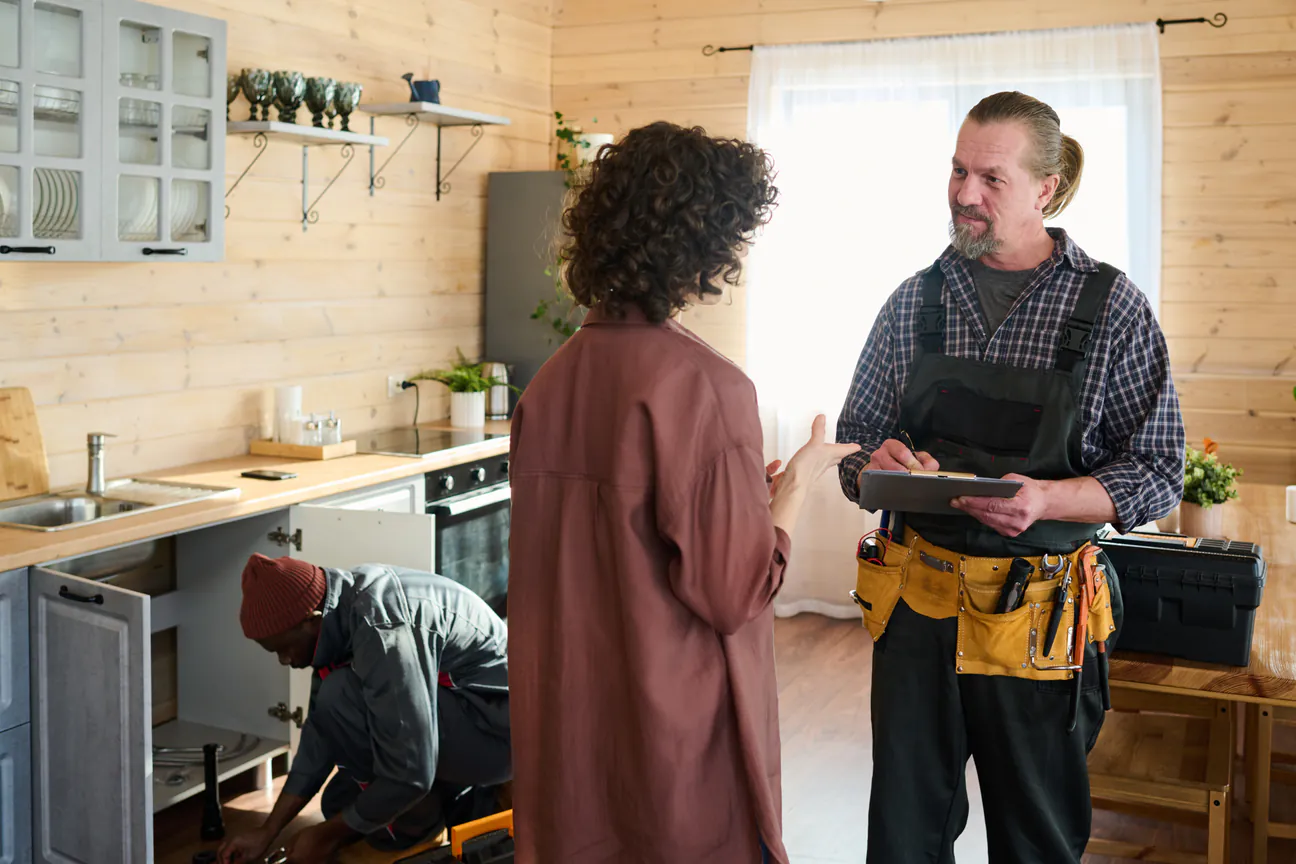 Repair technician taking notes on a clipboard while a homeowner explains the job in her kitchen, a second technician working under the sink