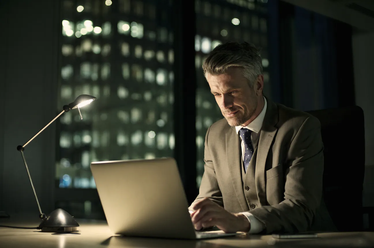 Lawyer in a suit working late on a laptop in a dark office, city lights in the windows behind him