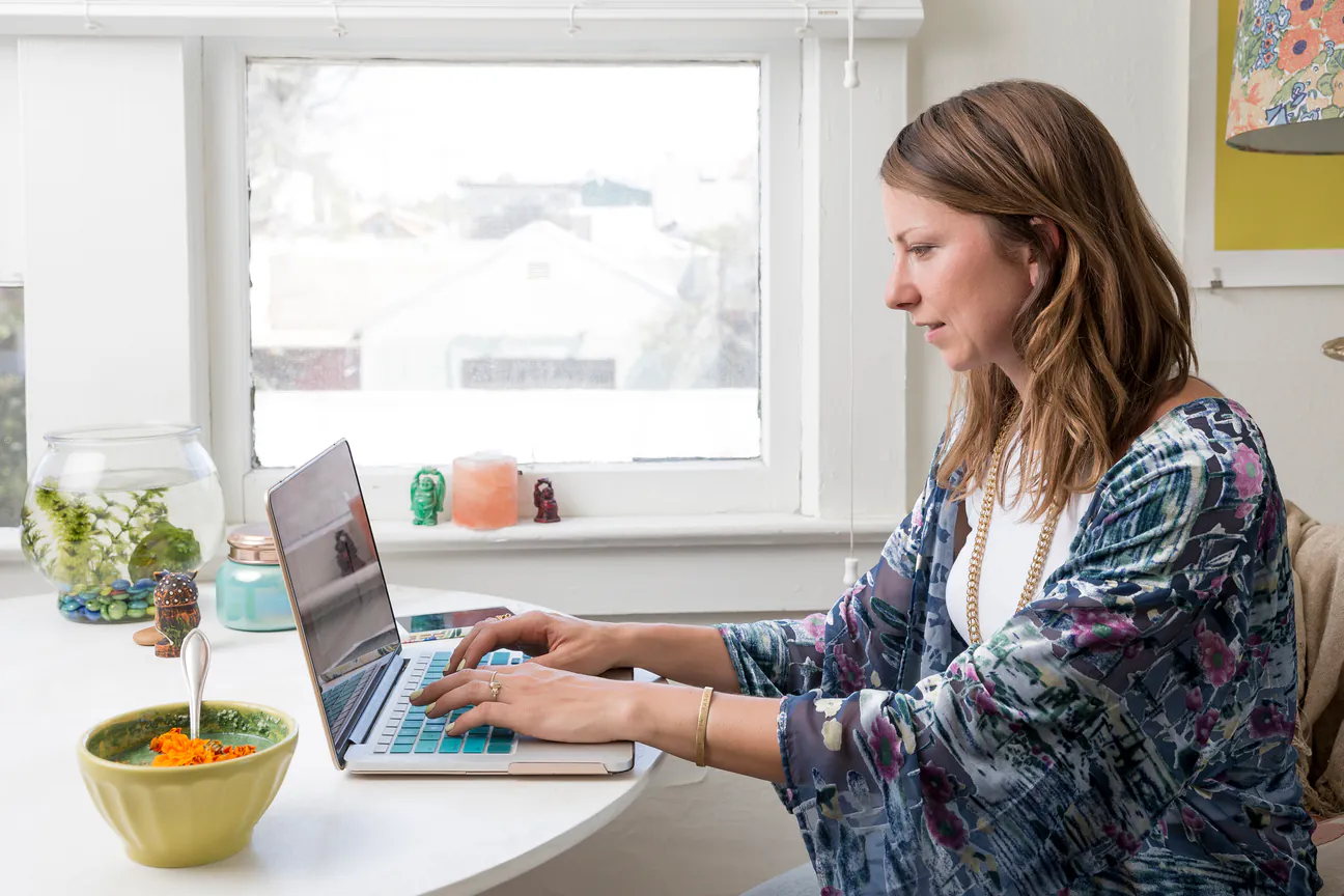 Woman types on a laptop at a round table by a window at home
